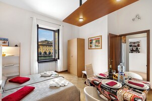 A hotel room with two beds, a dining table set for two, a window with a view of buildings, and a wardrobe.