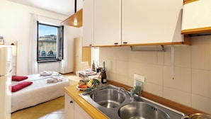 A compact hotel room with a kitchenette, a bed with red pillows, a window with a city view, and a sink with a wine bottle on the counter.