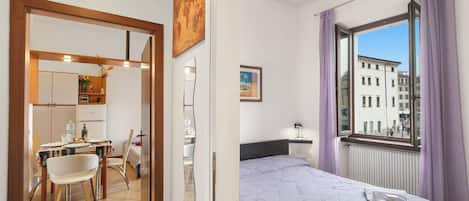 Ein Hotelzimmer mit Bett, Fenster mit Vorhängen, Blick auf Gebäude, Essbereich mit Tisch und Stühlen sowie Küchenzeile.