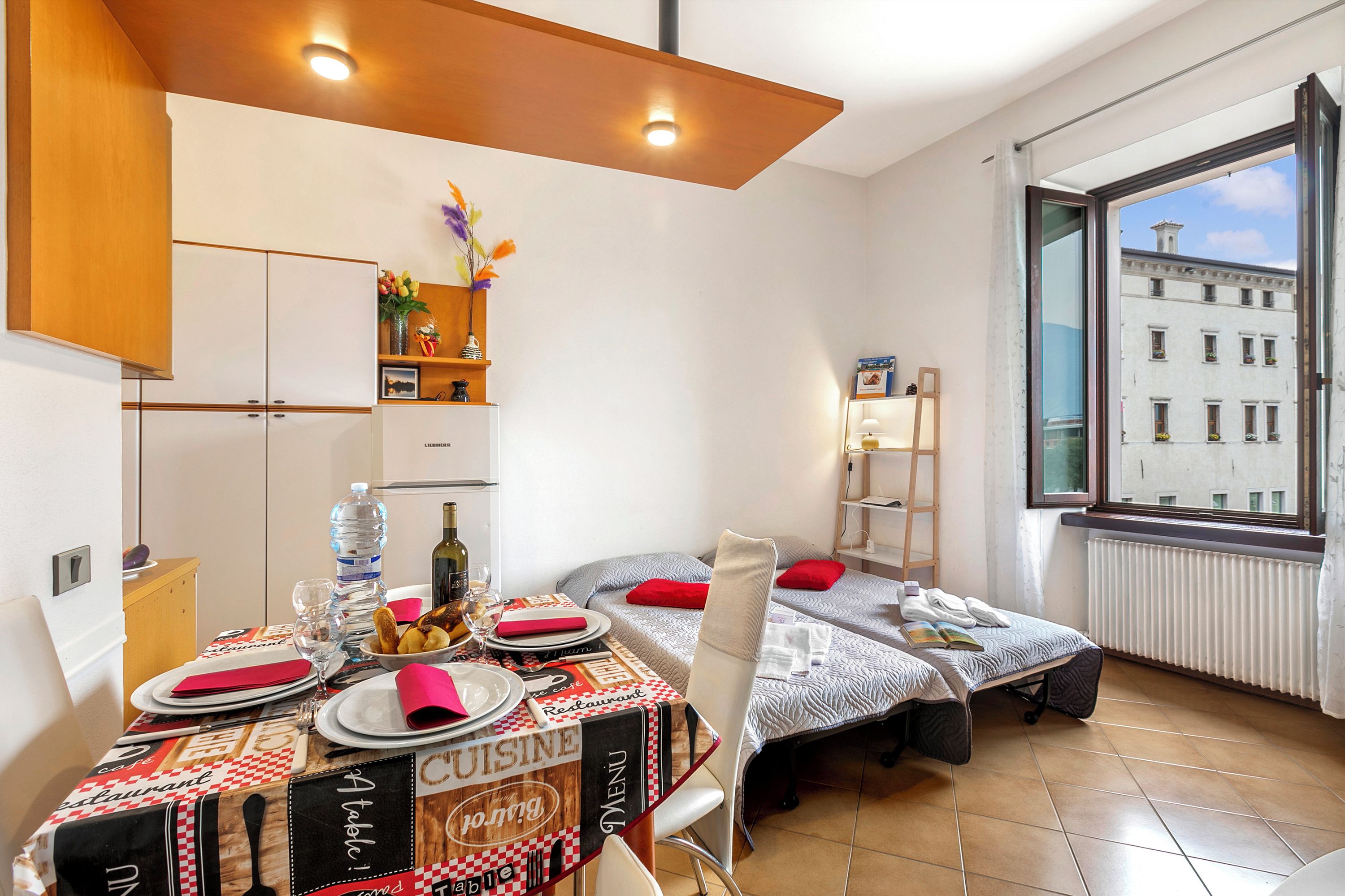 Un coin repas avec une table dressée pour deux personnes, un espace cuisine avec des placards et une fenêtre donnant sur des immeubles.