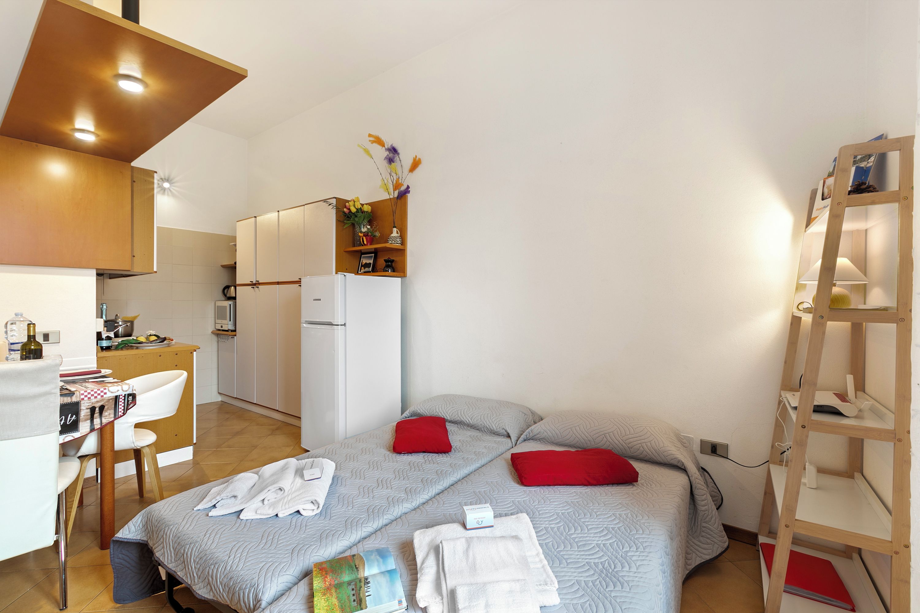 Une chambre d’hôtel compacte comprenant un coin cuisine, un lit avec des oreillers rouges, un bureau avec une chaise et une échelle en bois.