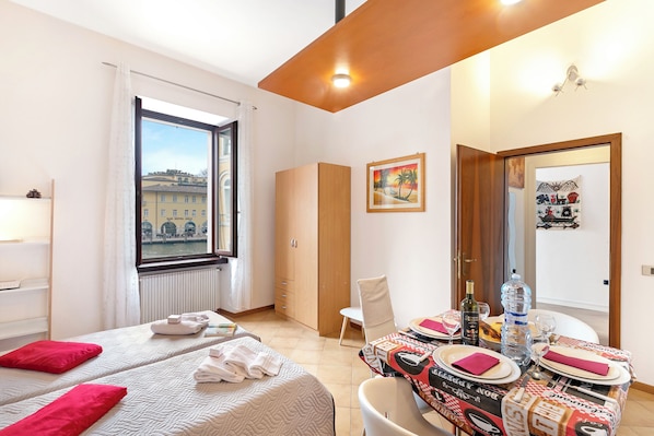 Une chambre d’hôtel avec deux lits, une table à manger dressée pour deux personnes, une fenêtre donnant sur la ville et une porte menant à une autre pièce.