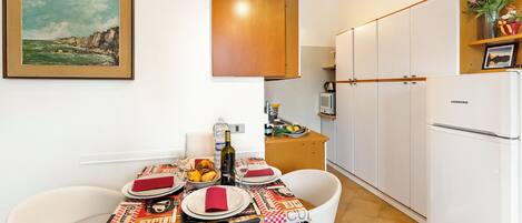 A dining area with a table set for four, a kitchen with white cabinets, and a framed painting on the wall.