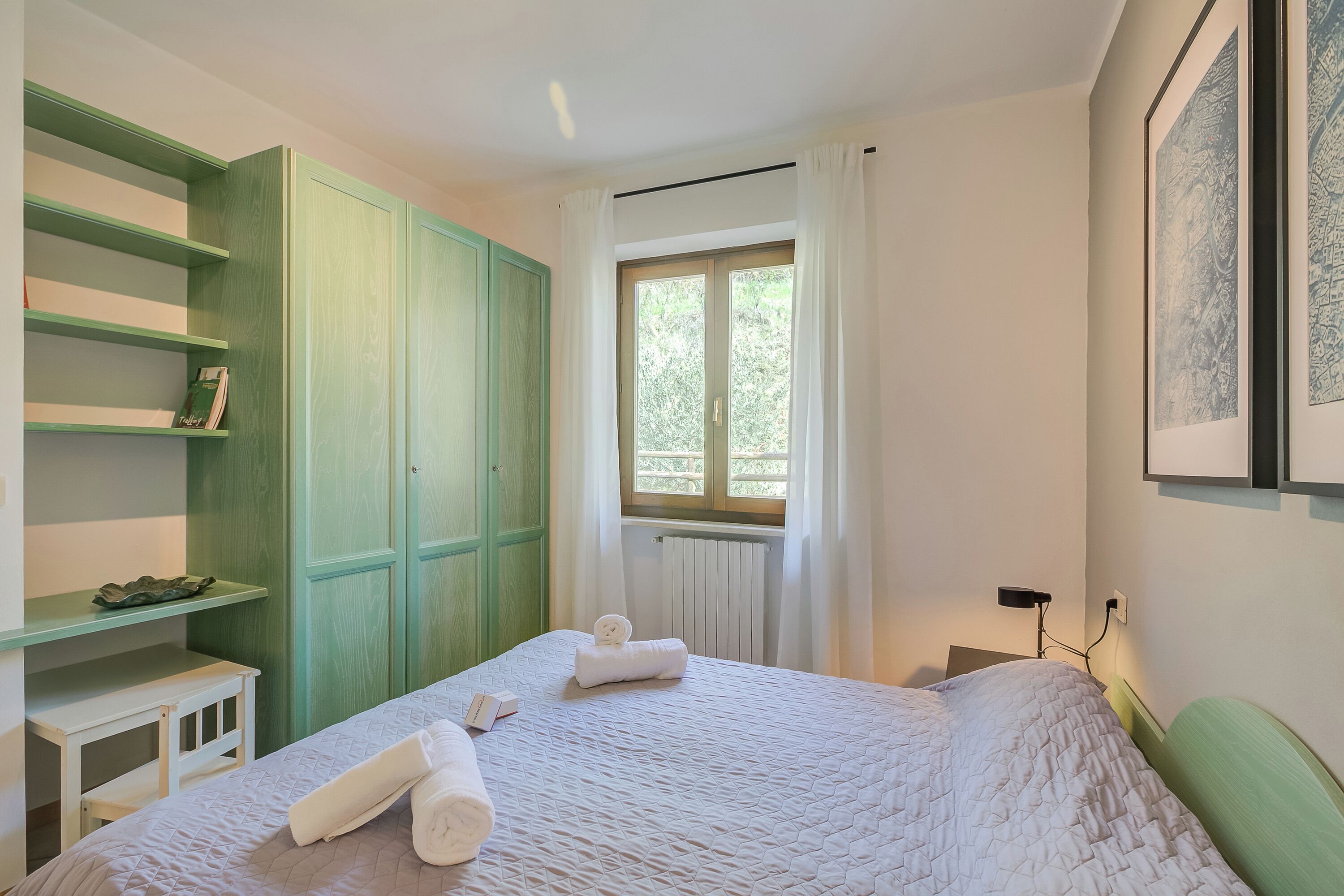 A bedroom with a bed, a green wardrobe, a small table, a window with curtains, and a framed picture on the wall.