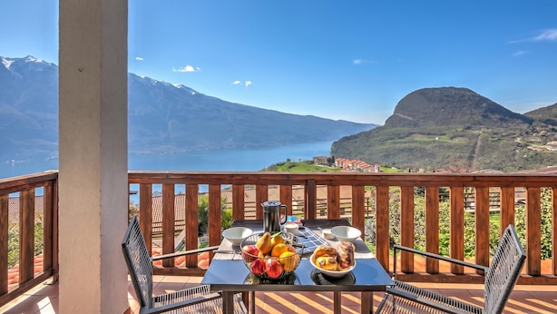 A balcony with a table set for breakfast, a view of mountains and a lake, and a wooden railing. - Balcone Sul Lago (Tremosine)