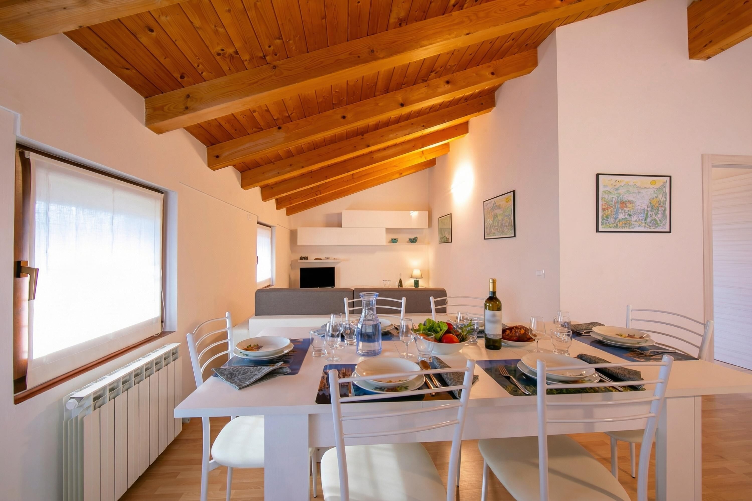 A dining area with a white table set for four, wooden ceiling beams, a radiator, and a window with blinds.