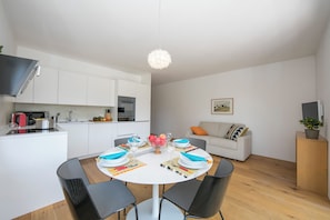 Una cocina moderna con armarios blancos, una mesa de comedor lista para cuatro personas y una sala de estar con un sofá y una televisión.