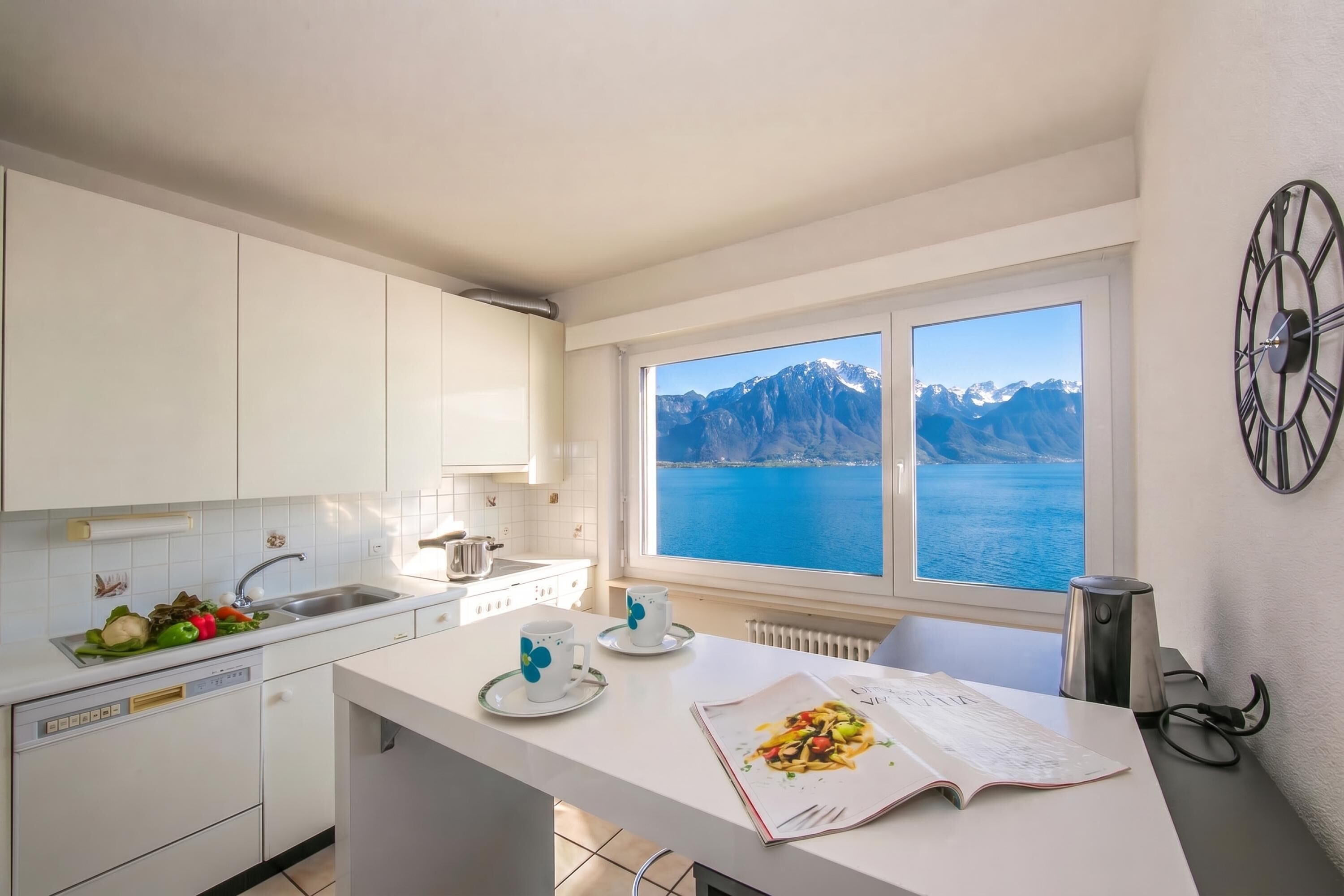 A modern kitchen with white cabinets, a large window with a mountain view, and a dining area with a magazine.