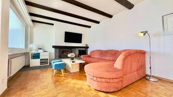 A modern living room with a wooden herringbone floor, a pink sofa, a coffee table, a blue ottoman, a fireplace, and a TV mounted on the wall.