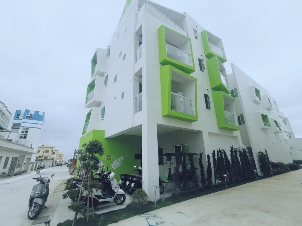 A modern multi-story residential building with balconies, a parking area, and a row of trees.