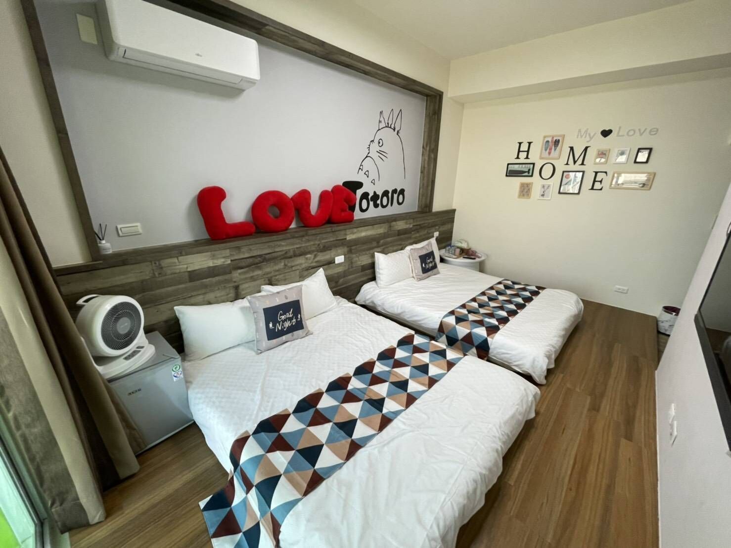 Habitación de hotel con dos camas, aire acondicionado mural, un pequeño ventilador y un cartel decorativo con la palabra 'LOVE' e ilustración de un gato.