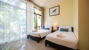 A hotel room with two beds, a large window with curtains, a framed picture on the wall, and a lamp on the nightstand.