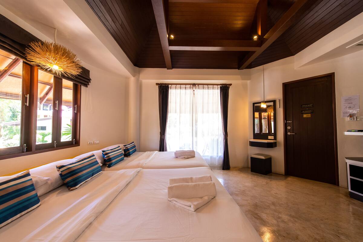 A hotel room with two beds, a wooden ceiling, a window with curtains, a door with a sign, and a small table with a lamp.