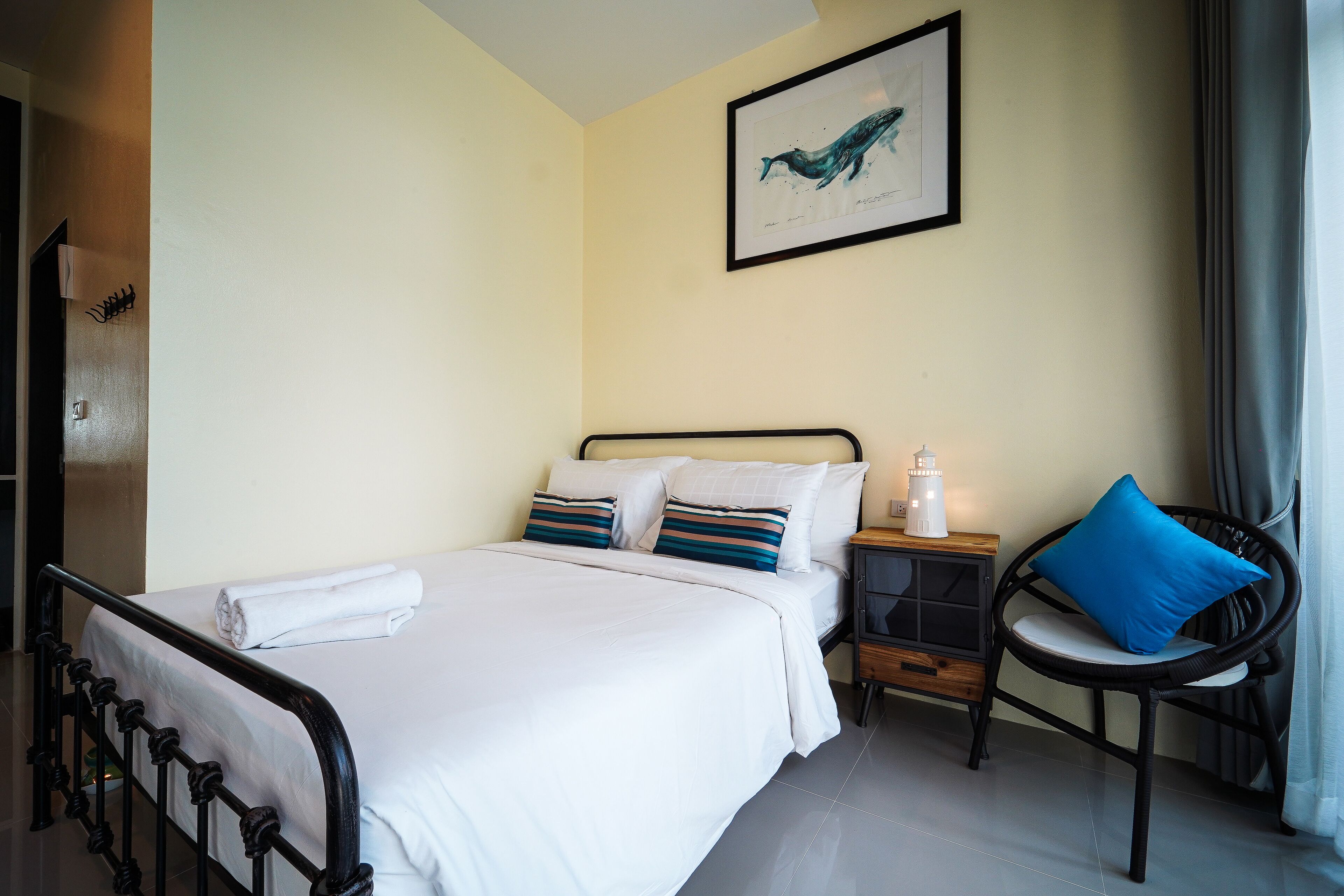 A neatly made bed with white linens and striped pillows, a black metal headboard, a bedside table with a lamp, and a blue cushion on a wicker chair.