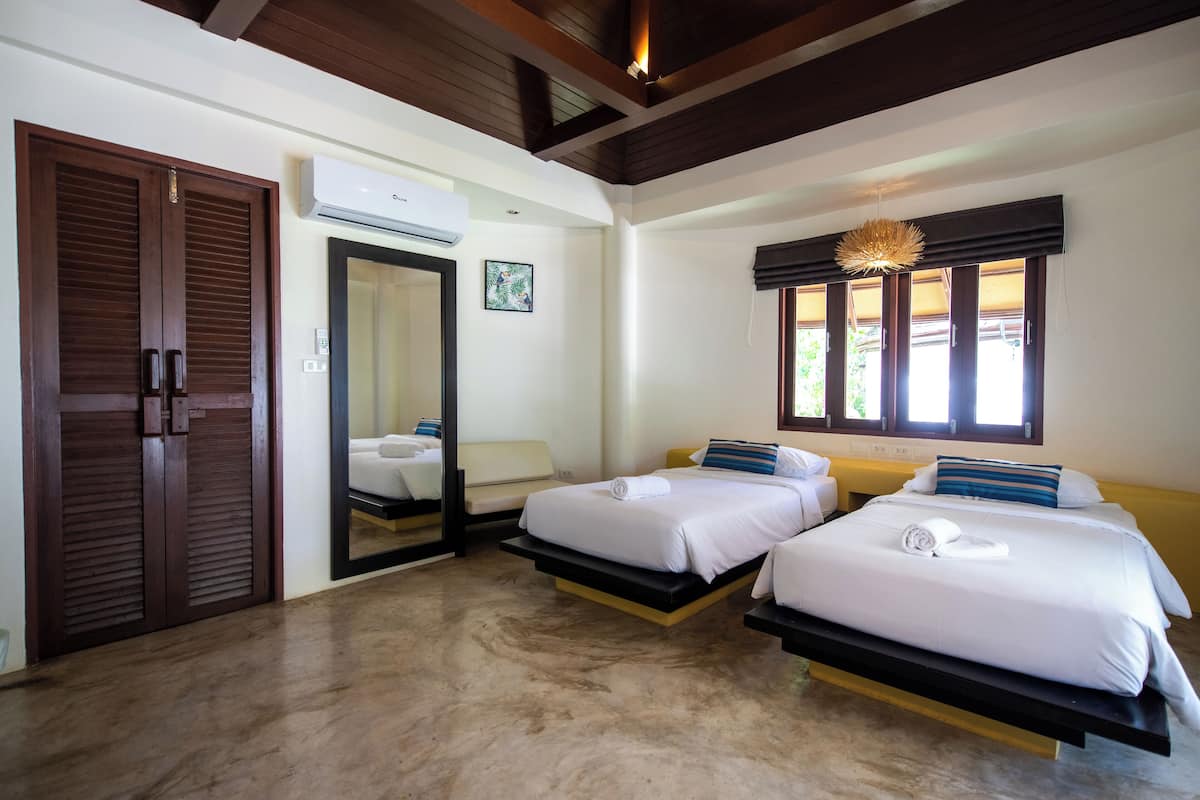 A hotel room with two beds, a wooden door, an air conditioning unit, and a window with a view of greenery.