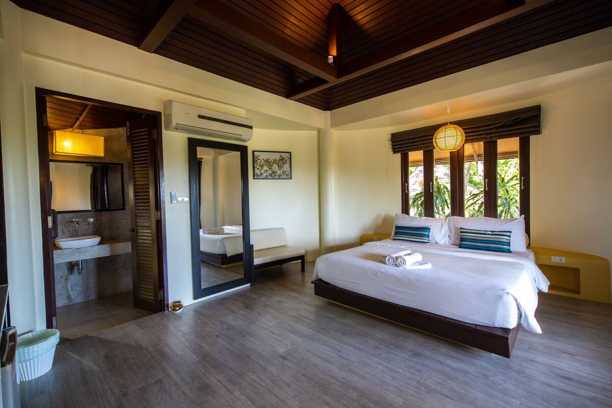 A bedroom with a large bed, a bedside table, a nightstand, a window with a view of greenery, and a bathroom with a sink and mirror.