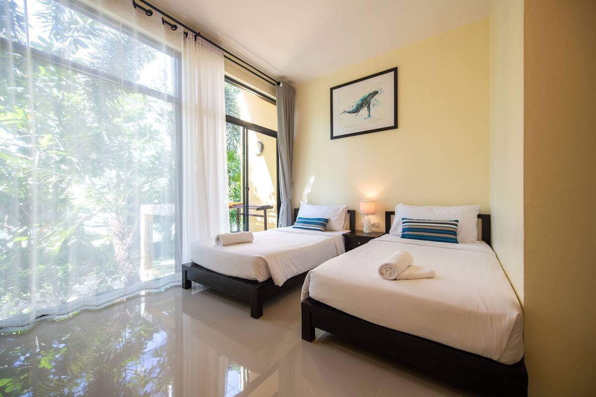 A hotel room with two beds, a window with curtains, a framed picture on the wall, and a nightstand with a lamp.