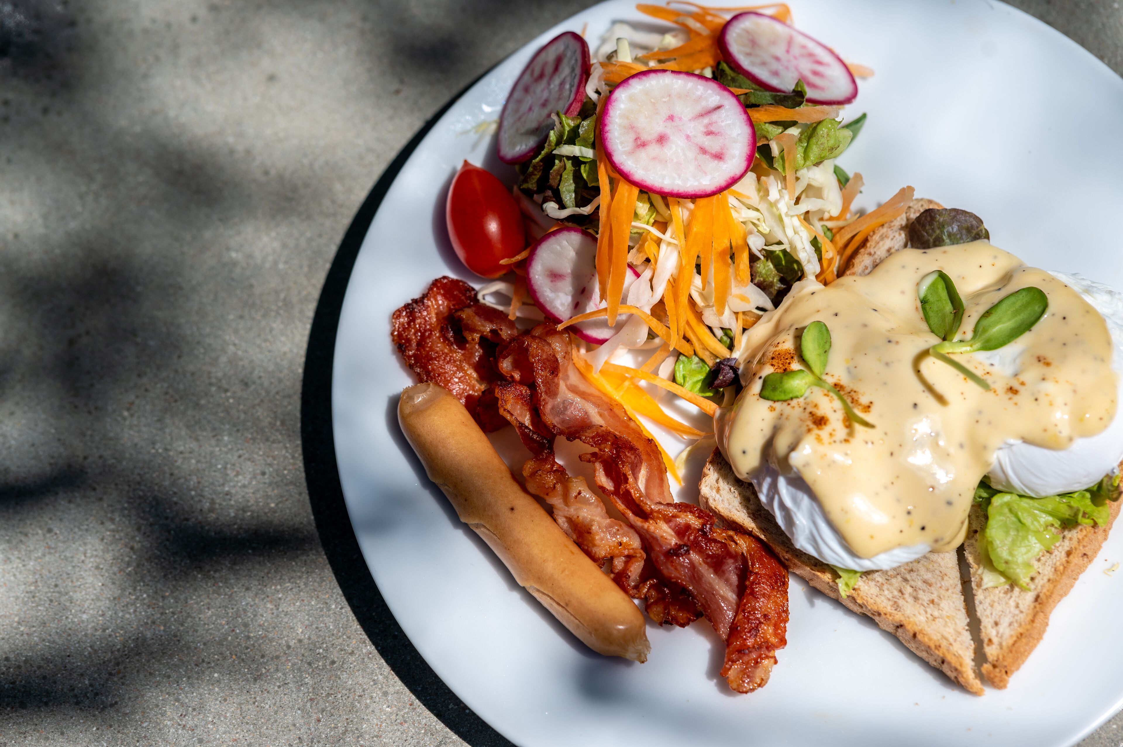 Een bord met een sandwich, bacon, een hotdog en een salade met radijsjes en geraspte wortelen.