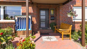A brick building with a covered porch, a bench, a striped chair, and a hanging flower basket. - Double room - Double room in the Hotel-Pension Marlies (Neuharlingersiel)