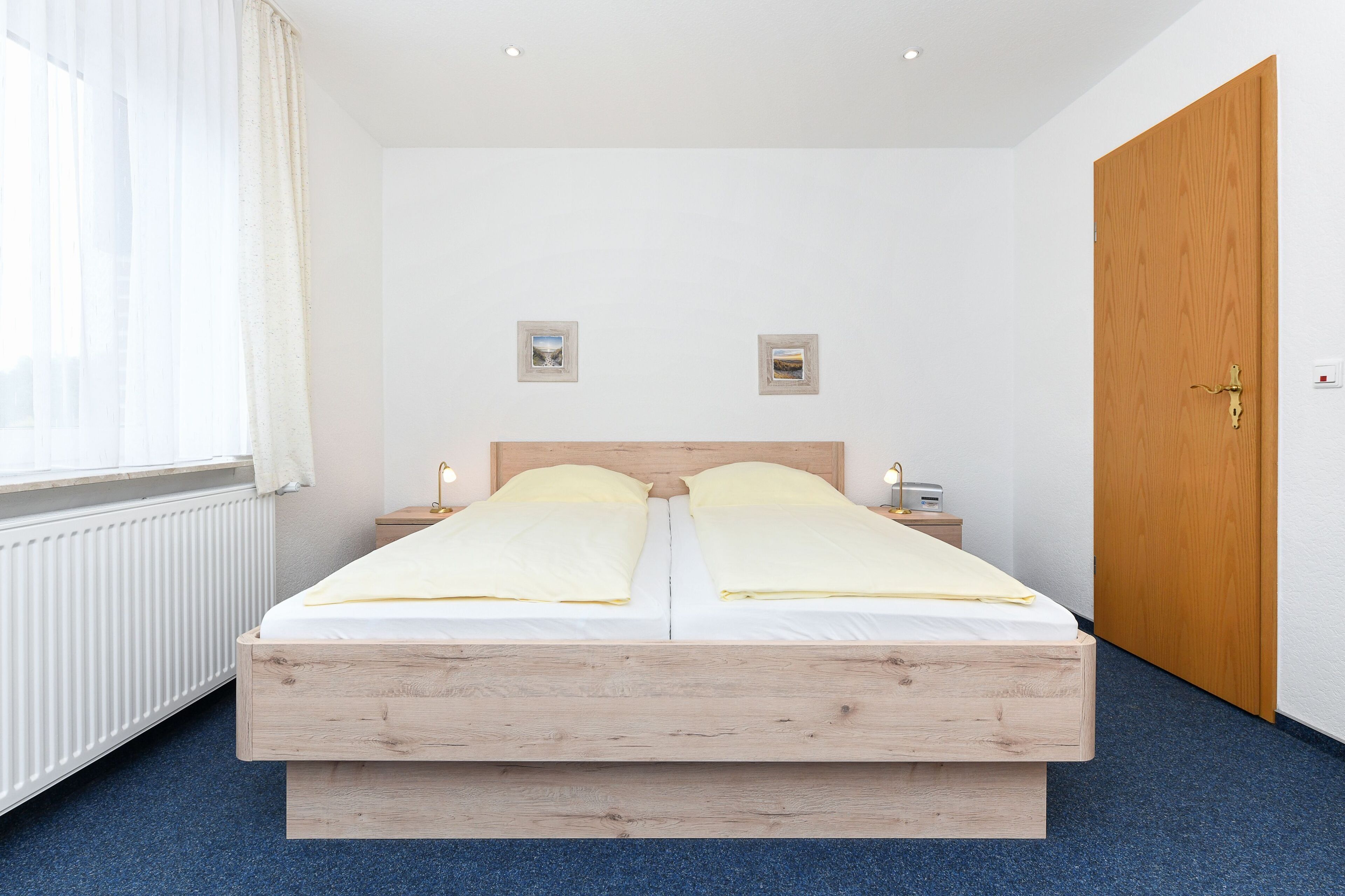 A double bed with wooden headboard, two bedside tables with lamps, a wooden door, and a window with sheer curtains.