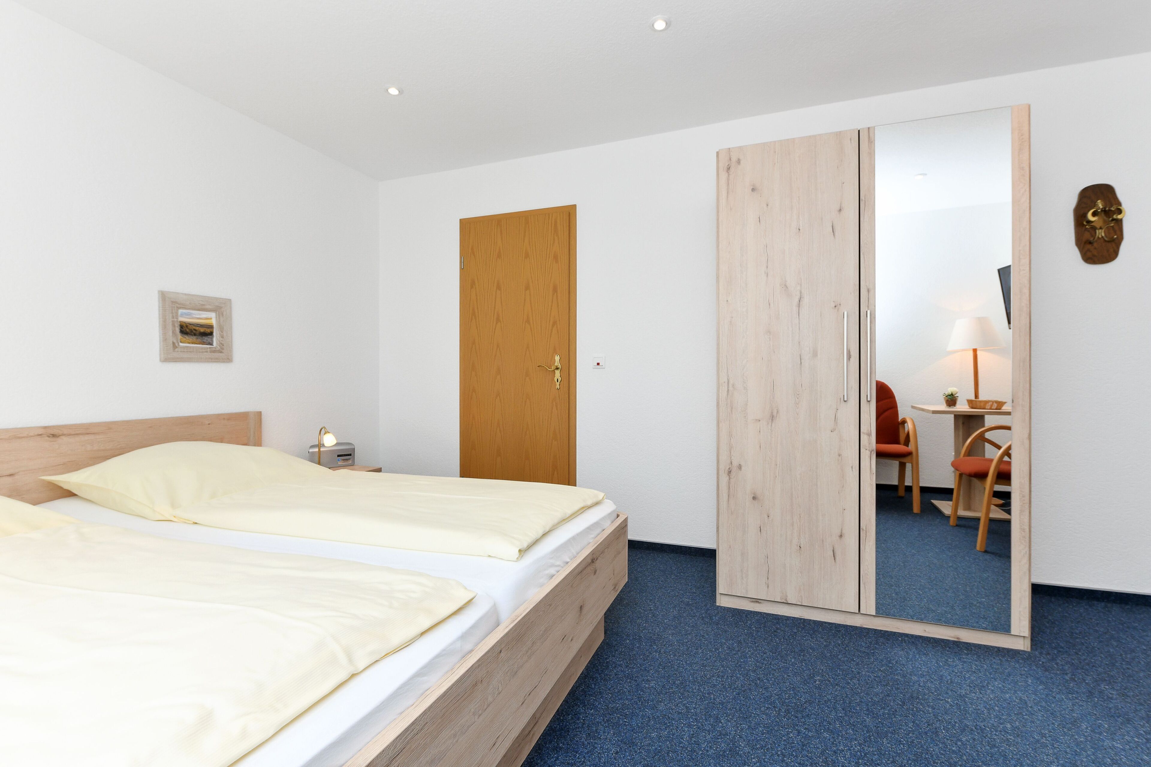 A hotel room with a wooden bed, a wardrobe, a desk with a chair, and a small table with a lamp.