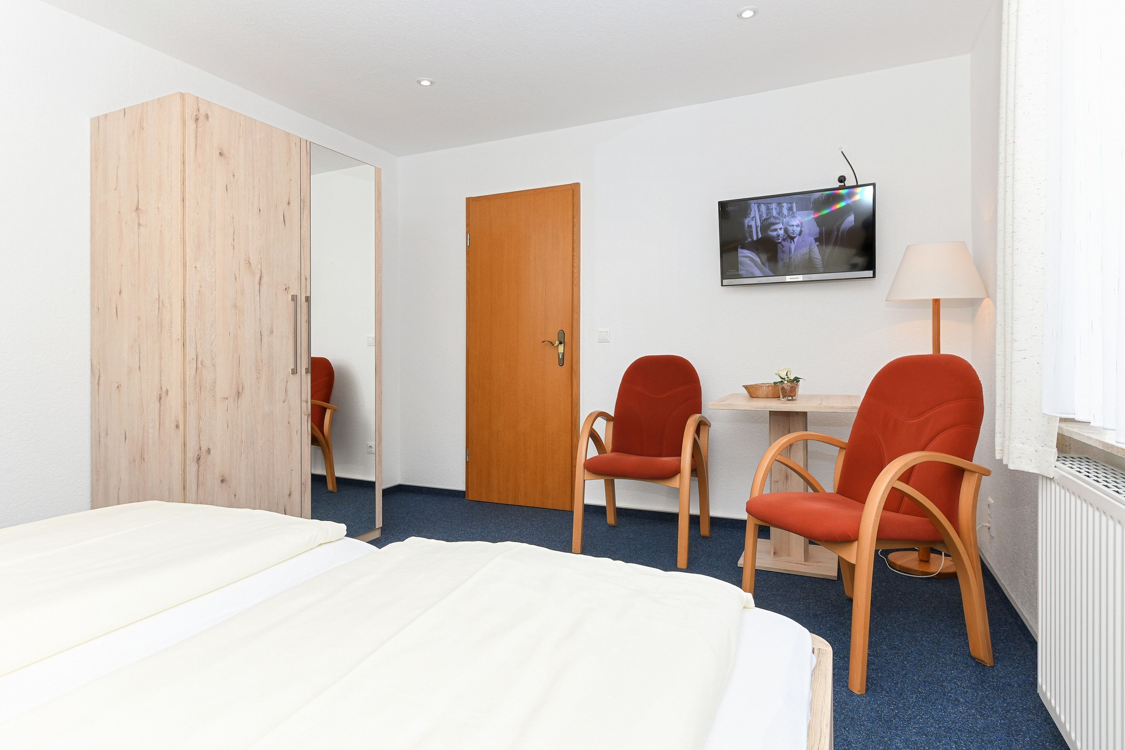 Een hotelkamer met een bed, twee rode stoelen, een houten garderobe, een bureau met een lamp en een televisie aan de muur.
