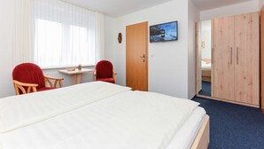 A hotel room with a bed, two red chairs, a small table, a wooden wardrobe, and a window with sheer curtains. - Double room - Double room in the Hotel-Pension Marlies (Neuharlingersiel)
