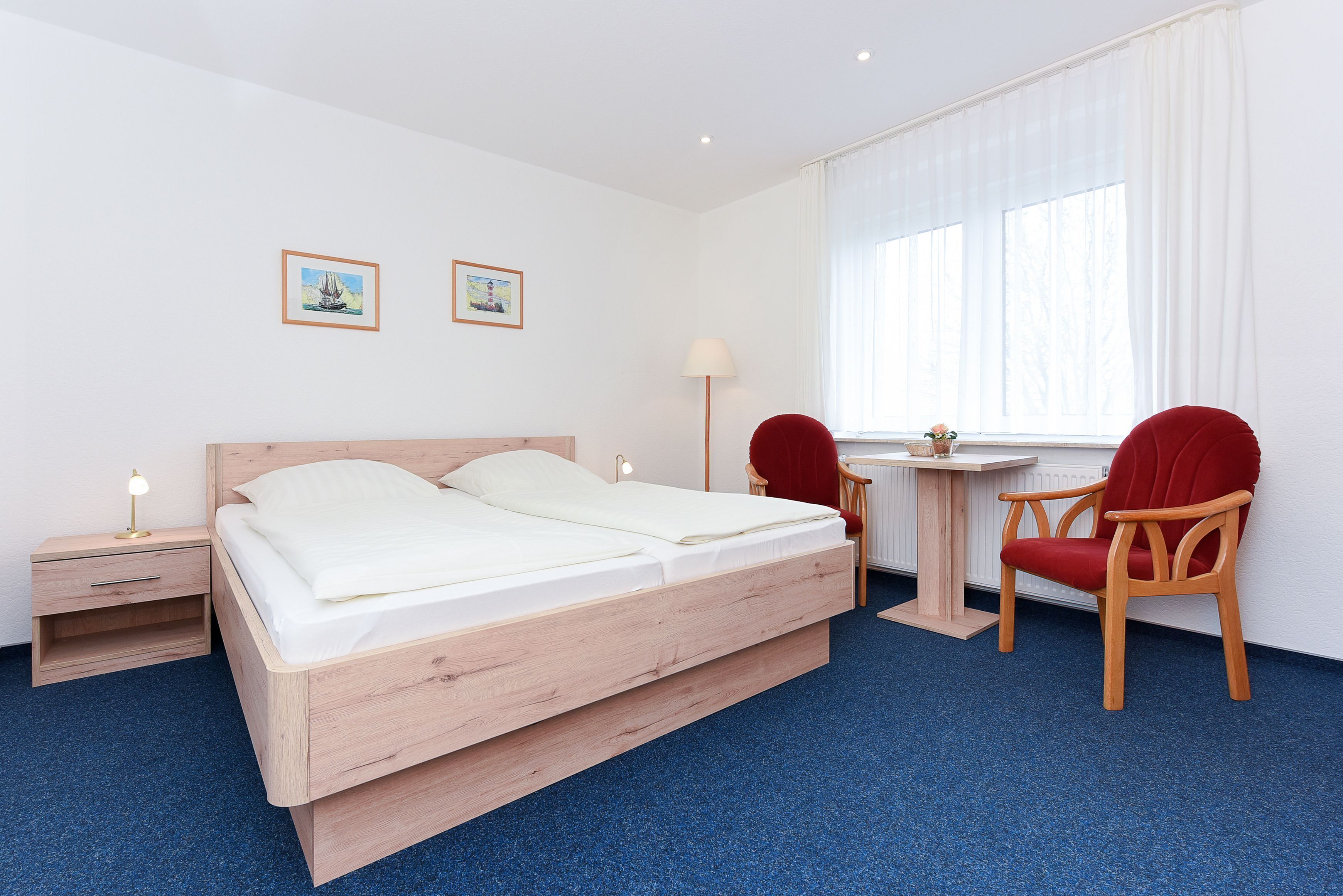 A hotel room with a wooden bed, a small desk, a chair, and two framed pictures on the wall.