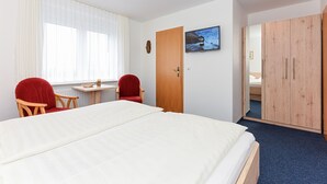 Habitación de hotel con cama, dos sillas rojas, una mesita, un armario de madera y una ventana con cortinas traslúcidas.