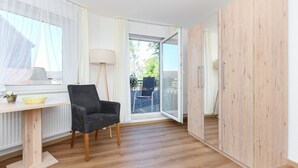 Ein Zimmer mit Holzboden, einem schwarzen Stuhl, einem kleinen Tisch mit einer gelben Schüssel, einer Lampe und einer Schiebetür zu einem Außenbereich mit Tisch und Stühlen.