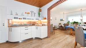 A modern kitchen with white cabinets, a wooden countertop, and a brick column. There is a dining area with a blue tablecloth and chairs.