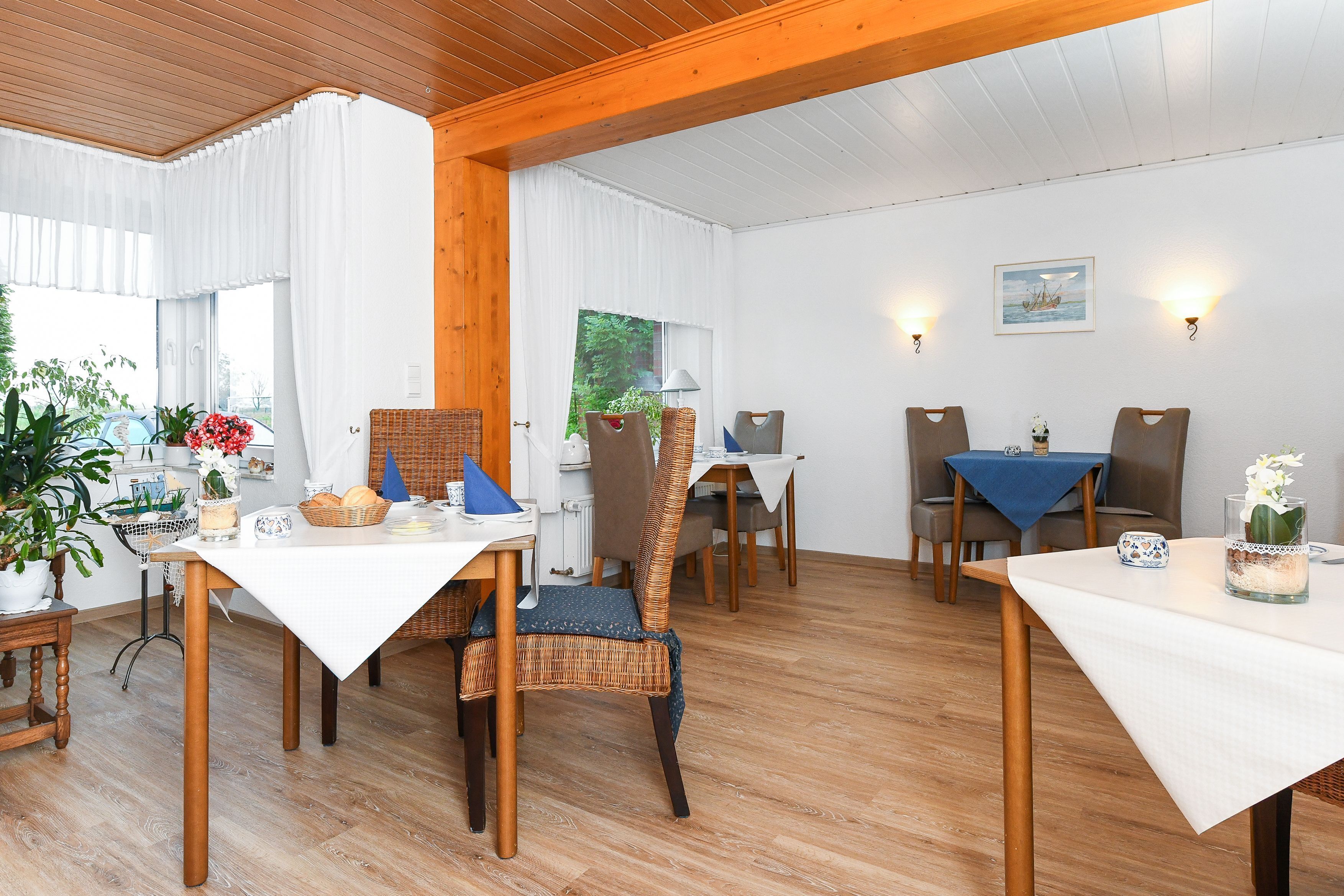 Zona pranzo con tavoli e sedie in legno, tovaglie bianche e piante in vaso.