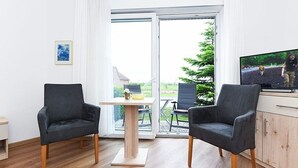 A modern room with two black chairs, a wooden table, a TV, and a sliding glass door leading to an outdoor area with a table and chairs.