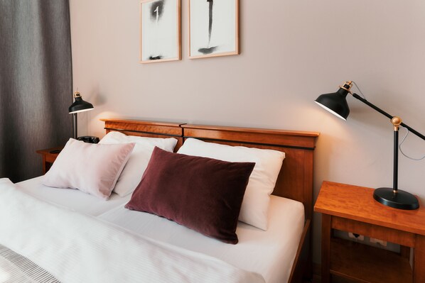 Una cama bien hecha con almohadas blancas y burdeos, un cabecero de madera y dos mesitas de noche con lámparas.