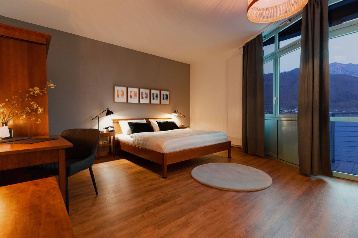A modern hotel room with a wooden bed, a desk, a chair, and a view of a body of water through large windows.