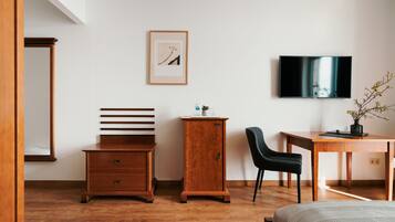Une chambre avec une commode en bois, un bureau avec une chaise, un lit, un téléviseur fixé au mur et un tableau encadré.