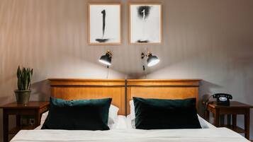 Habitación de hotel con cabecera de madera, dos lámparas de noche, cama con ropa blanca, dos almohadas verdes, una planta en maceta y un teléfono sobre una mesita de noche de madera.