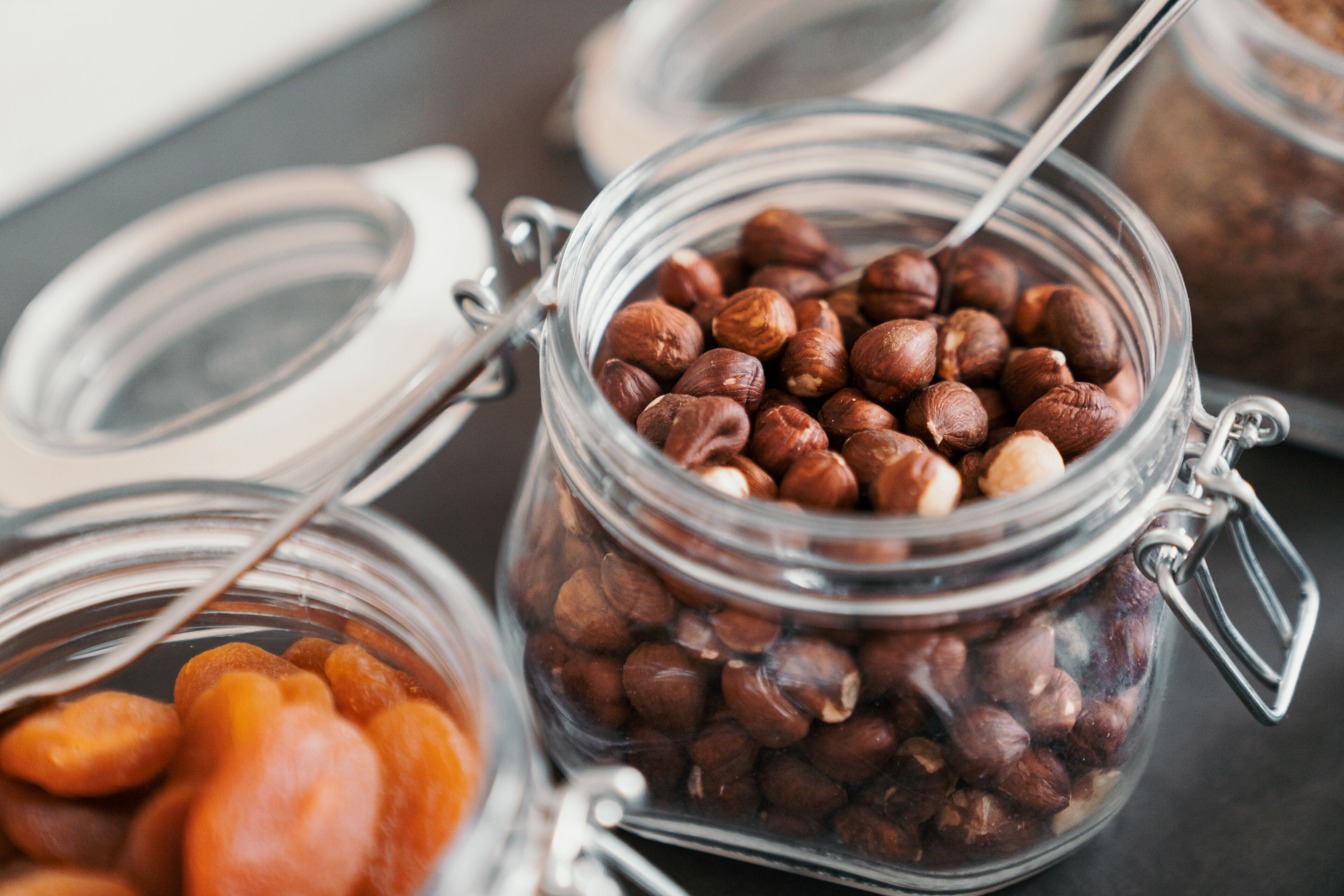 A glass jar filled with hazelnuts, a spoon inside, and a partially visible jar with a metal clasp lid.