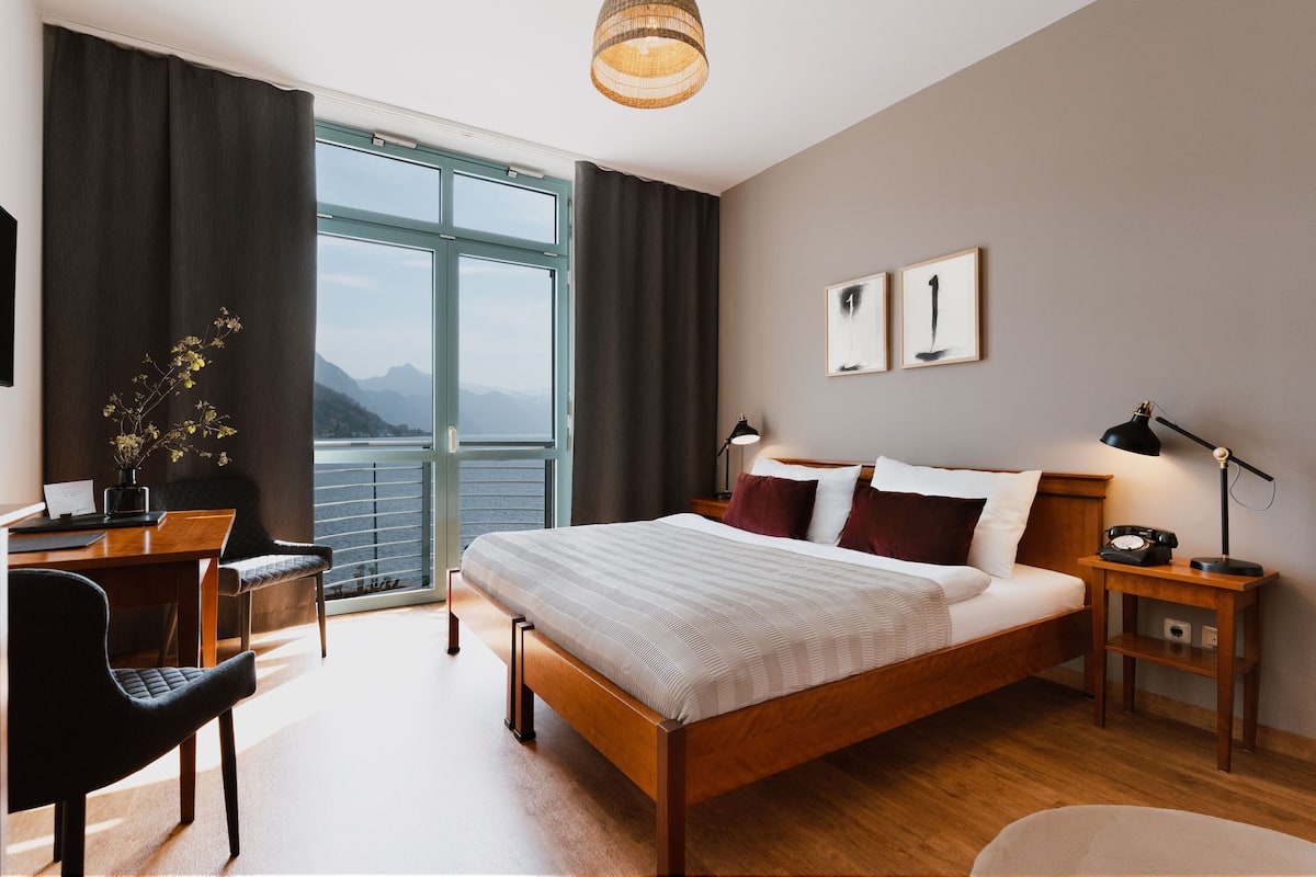 A modern hotel room with a large bed, a desk, a chair, a nightstand, and a view of mountains through the window.