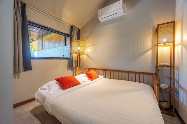 A bedroom with a bed, an air conditioner, a window with blinds, and a wall-mounted lamp.