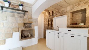 Une cuisine aux murs en pierre, aux armoires blanches et dotée d’une étagère intégrée où sont disposées des plantes.