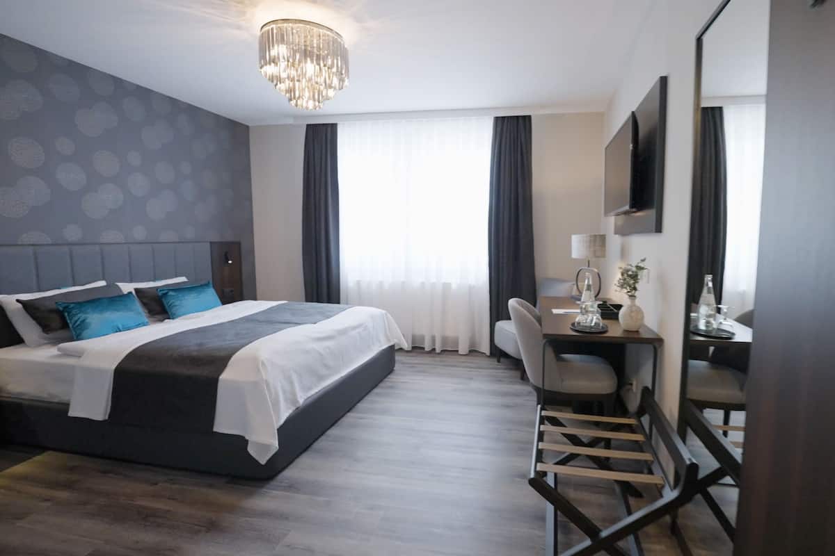 Una habitación de hotel moderna con una cama grande, un escritorio y una silla. Se puede ver una araña, un televisor y una ventana con cortinas.