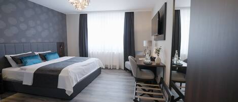 A modern hotel room with a large bed, a desk, and a chair. There is a chandelier, a TV, and a window with curtains.