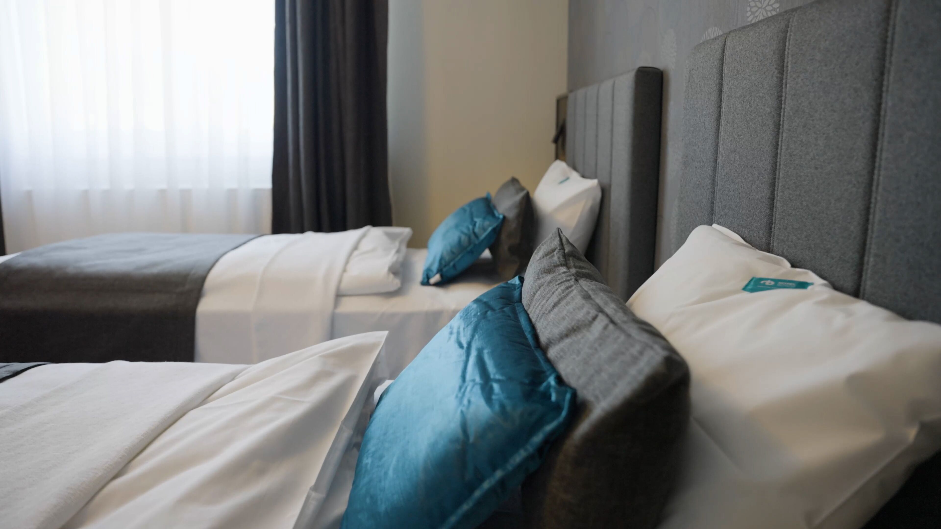 Ein Hotelzimmer mit zwei Betten, jeweils mit grauem Kopfteil und weißer Bettwäsche. Auf den Betten liegen blaue und graue Kissen.