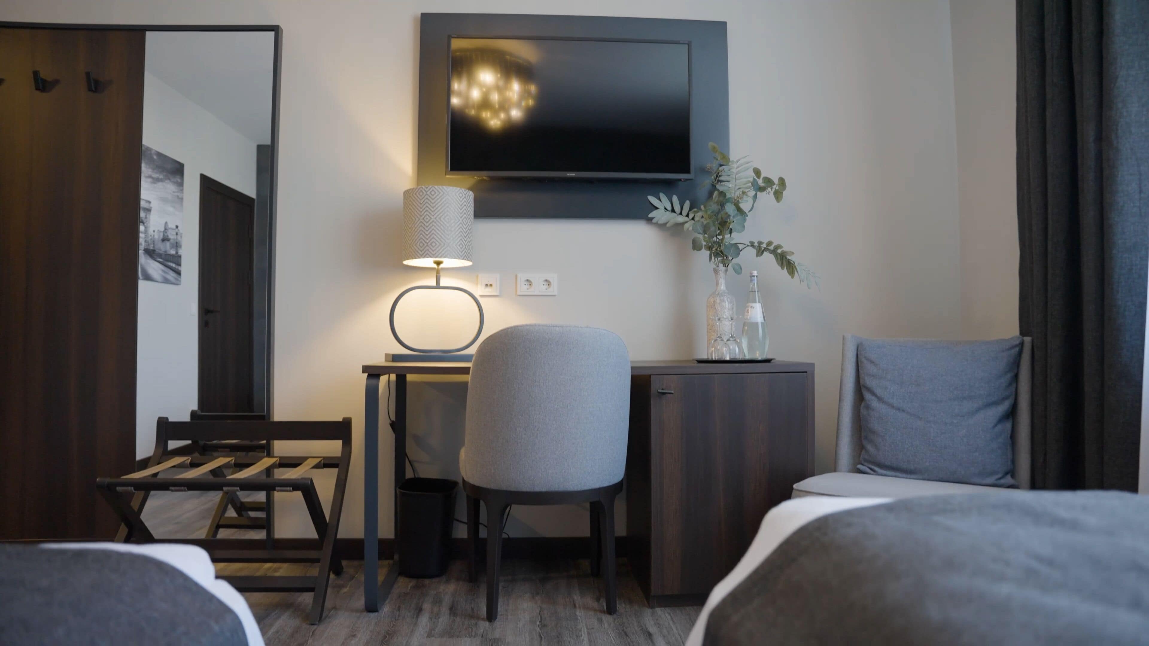 A hotel room with a bed, a desk, a chair, a TV, a lamp, and a small cabinet with decorative items.