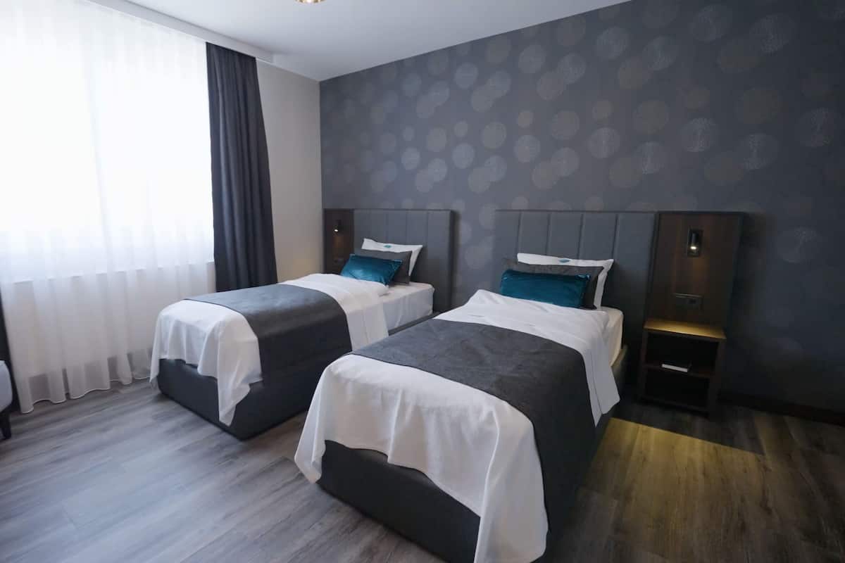 Habitación de hotel con dos camas, una silla, una mesita de noche y un ventanal con cortinas.