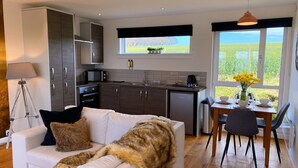 A modern kitchen with dark cabinets, a microwave, and a coffee maker. There is a dining area with a table set for two, and a sofa with a fur throw.
