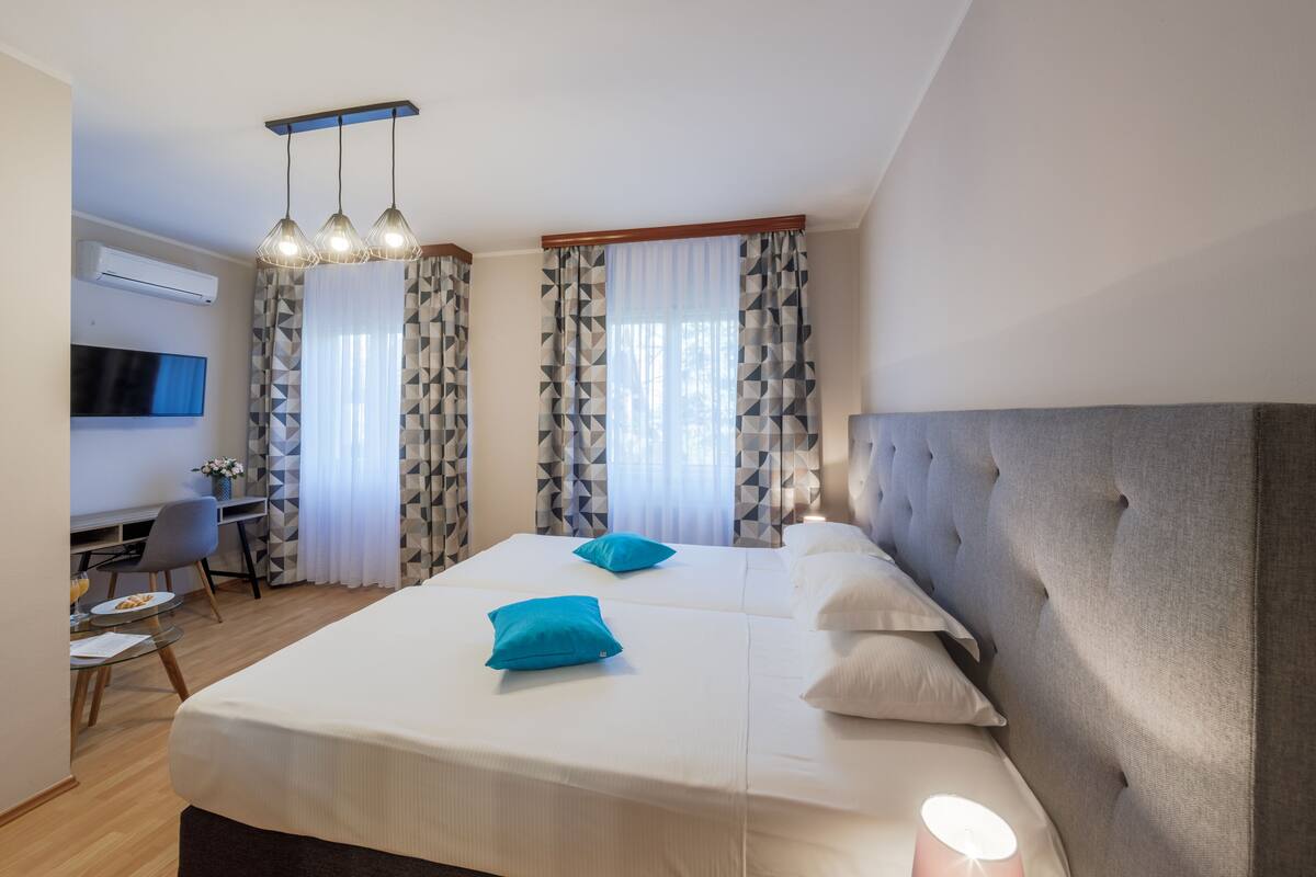 A hotel room with a large bed, two pillows, a desk with a chair, a TV, and a window with patterned curtains.