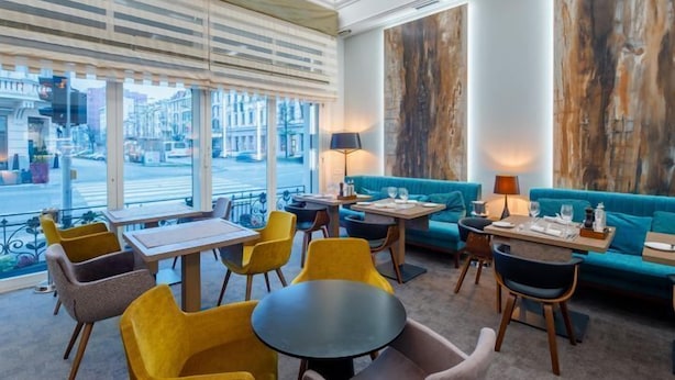 Un'area lounge moderna con divano, tavolini e sedie, ampie finestre con tende e vista sulla strada.