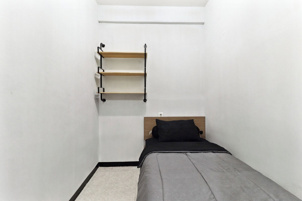 A small, single-bed room with a wooden headboard, a black pillow, and a wall-mounted shelf with three compartments.